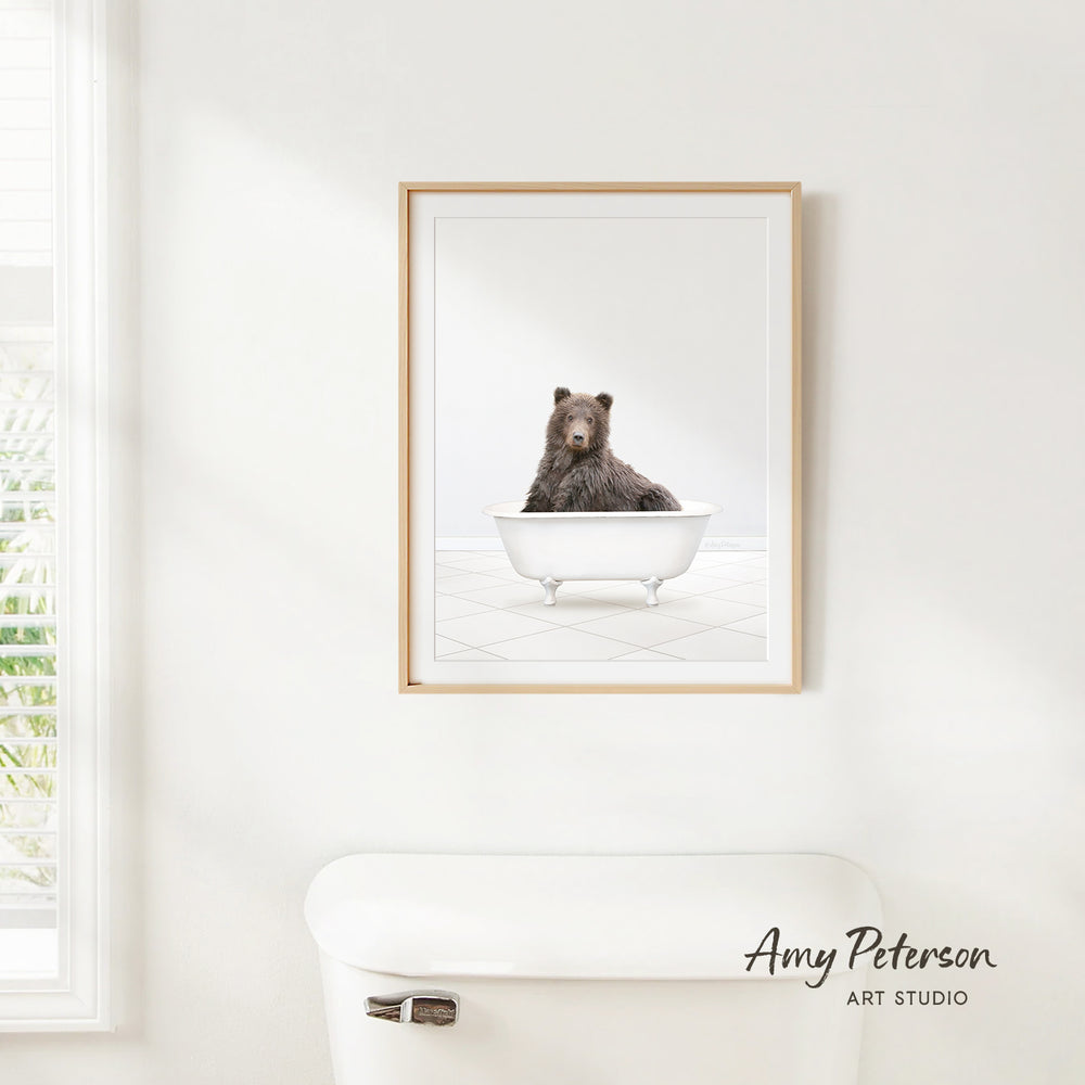 A framed image of a bear sitting in a white bathtub is hanging on a white wall.
