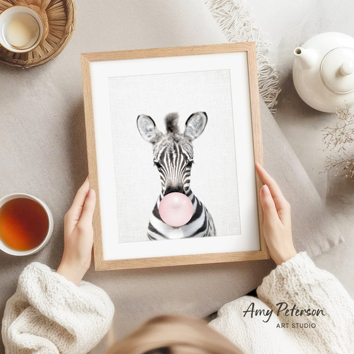 A person is holding a framed picture of a zebra blowing a bubble with a pink bubble gum bubble, with a teapot and a cup of tea nearby.