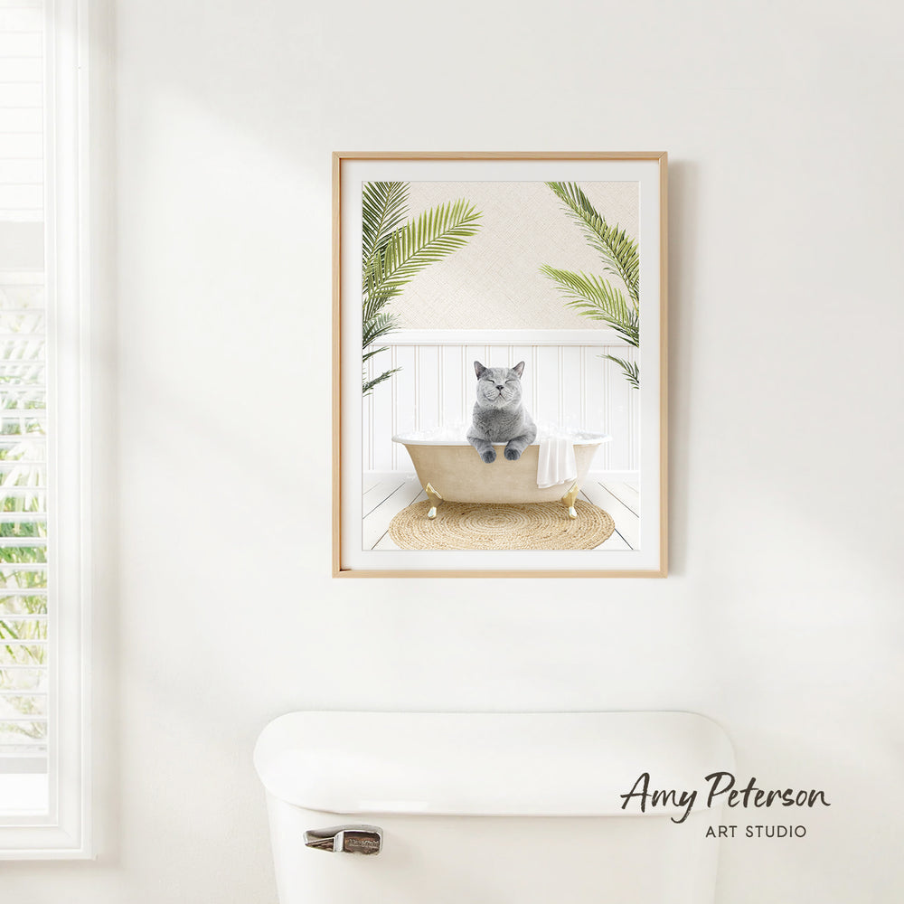 A framed image of a gray cat sitting in a bathtub, surrounded by green plants, is hanging on a white wall in a bathroom.