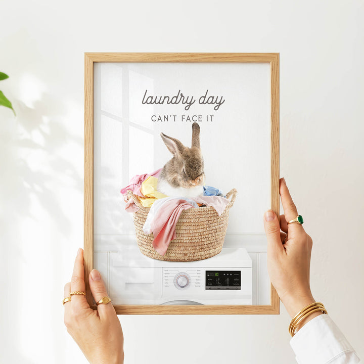 A person's hands are holding a framed picture of a rabbit sitting in a basket filled with laundry, with the text "laundry day can't face it" displayed on the picture.