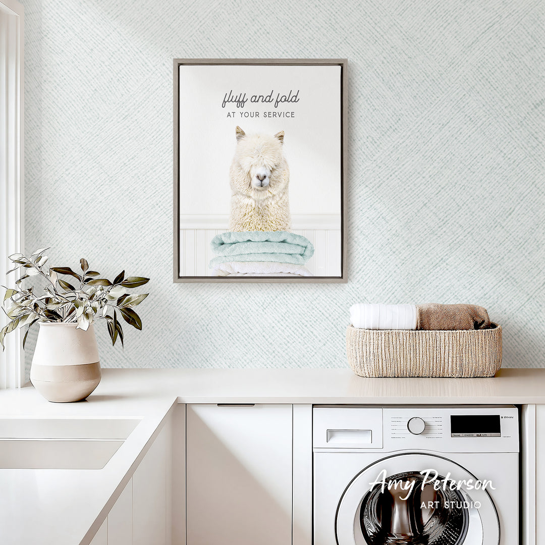 a framed picture of a fluffy alpaca sitting on a towel, hanging on a wall above a laundry basket and a white sink.