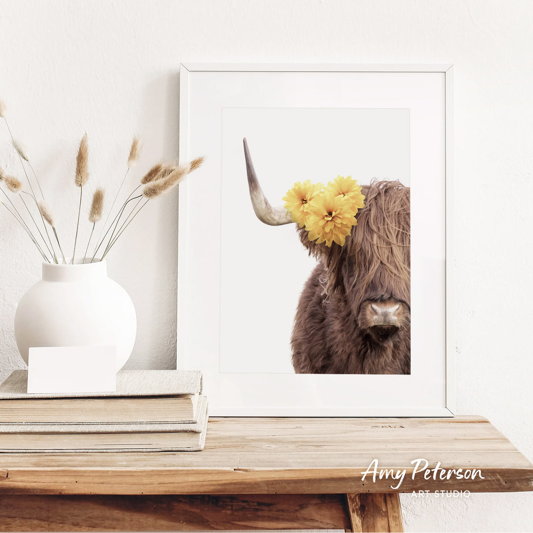 a framed picture of a cow with yellow flowers on its horns, placed on a wooden shelf next to a white vase with dried flowers.