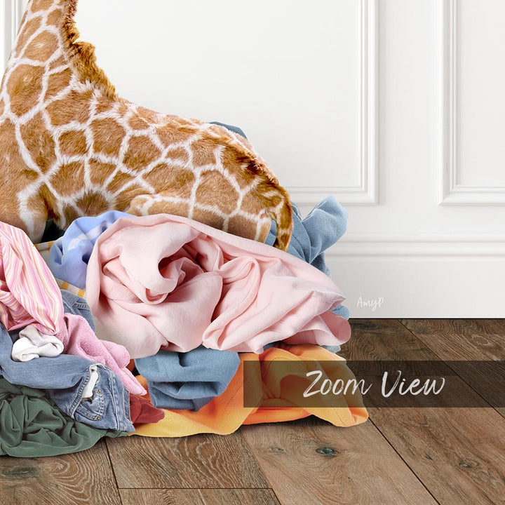 A giraffe standing on a pile of clothes, with a white wall and wooden floor in the background.