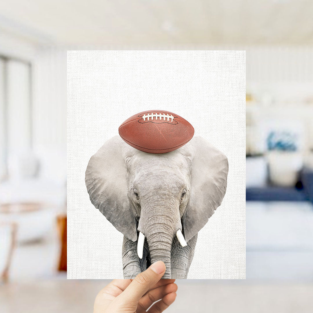 A hand holds up a framed image of an elephant with a football on its head.