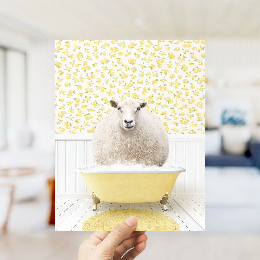 A hand holds up a framed image of a fluffy white sheep sitting in a yellow bathtub, surrounded by a yellow floral pattern on the wall.