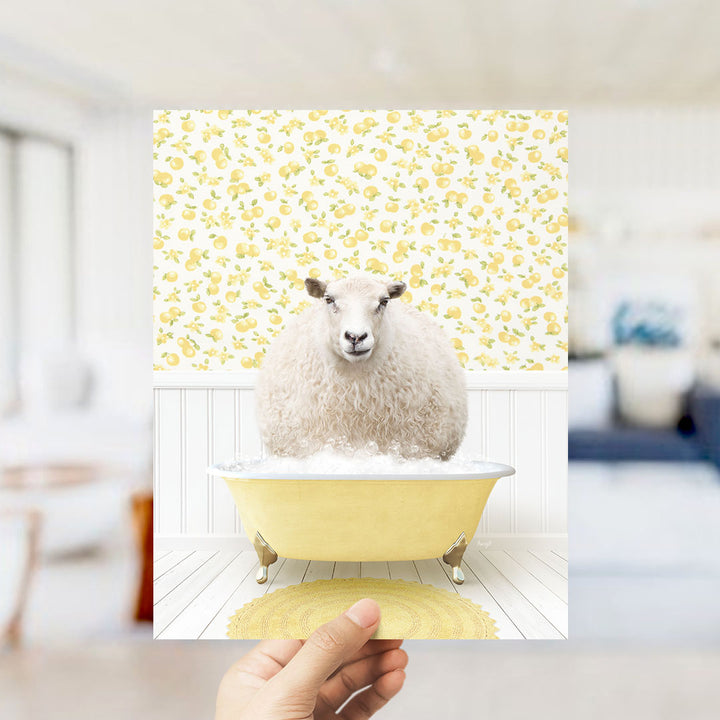 A hand holds up a framed image of a fluffy white sheep sitting in a yellow bathtub, surrounded by a yellow floral pattern on the wall.