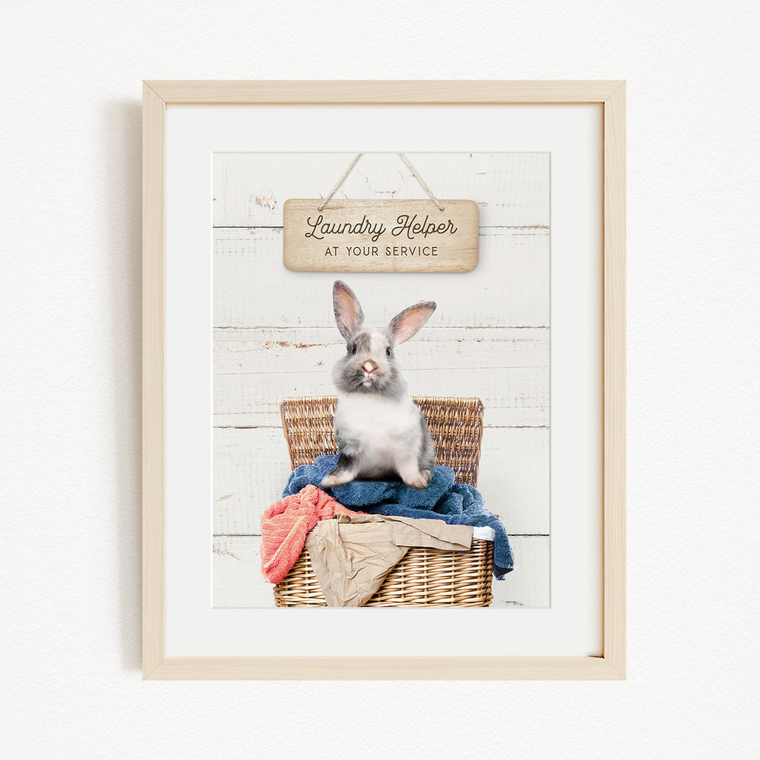 A framed image of a rabbit sitting in a wicker basket filled with clothes, with a sign above that reads "Laundry Helper at your service".