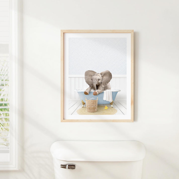 A framed image of a baby elephant sitting in a bathtub, surrounded by various objects such as a basket and a towel.