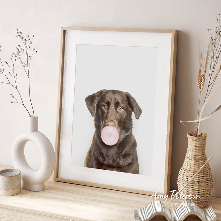 A framed photograph of a brown dog blowing a bubble gum bubble is displayed on a wooden shelf, accompanied by decorative vases and a woven basket.