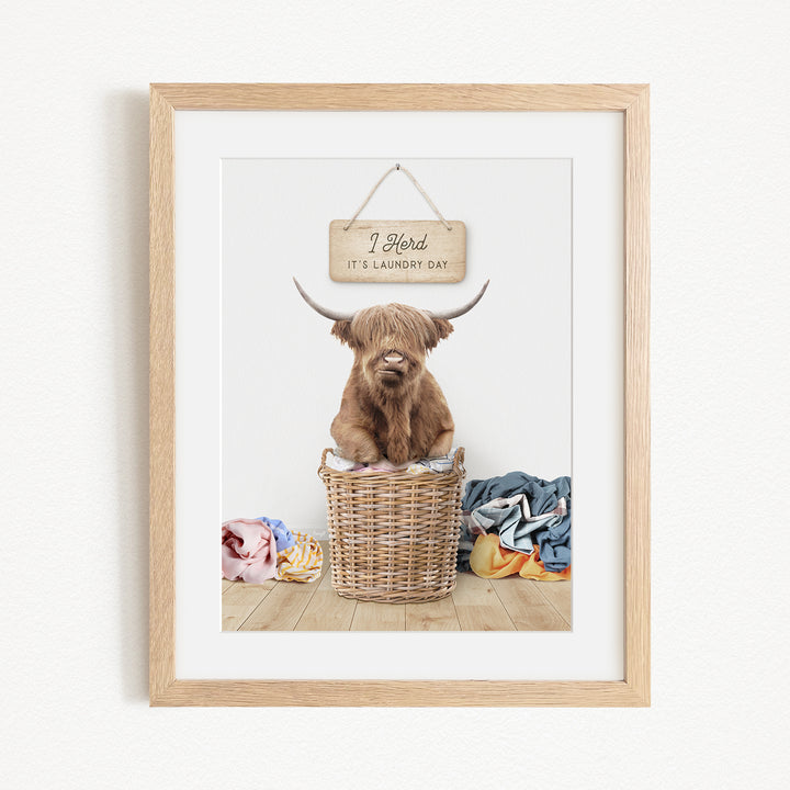 A framed image depicting a Highland cow sitting in a wicker basket, with a sign above it that says "I herd it's laundry day".