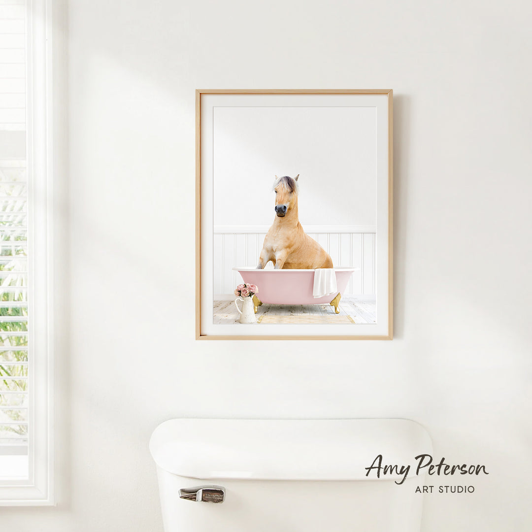 A framed photograph of a horse sitting in a pink bathtub, with a vase of flowers and a white toilet in the foreground.