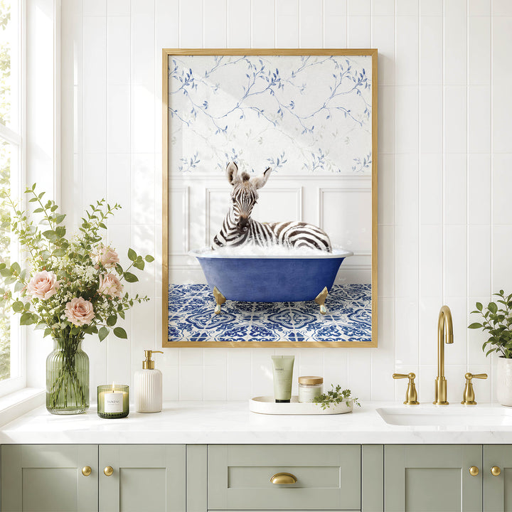 A framed image of a zebra sitting in a blue bathtub, surrounded by flowers and a decorative vase, is displayed on a white tiled wall above a bathroom counter with a sink and gold faucets.