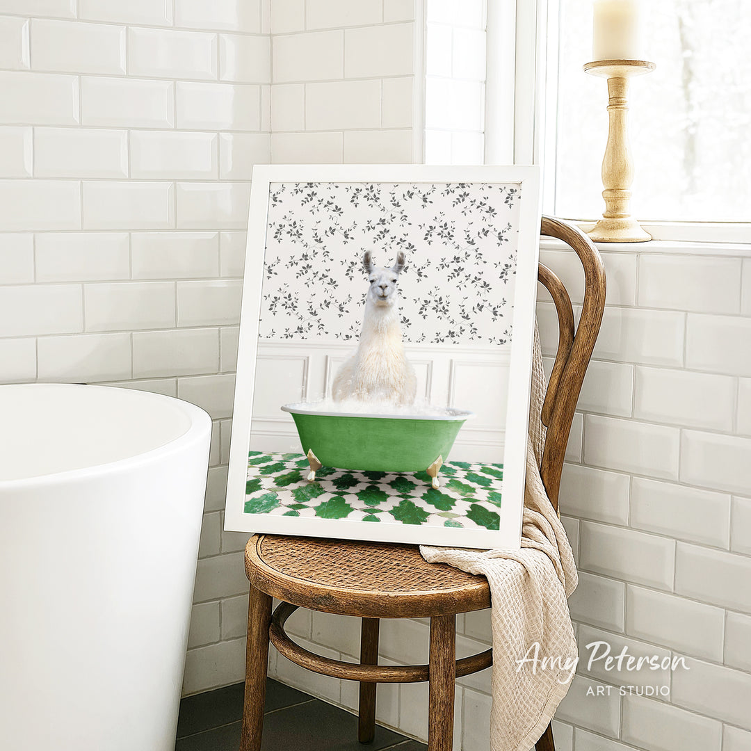 A framed image of a white llama sitting in a green bathtub, surrounded by a patterned design, is displayed on a wooden chair in a bathroom setting.
