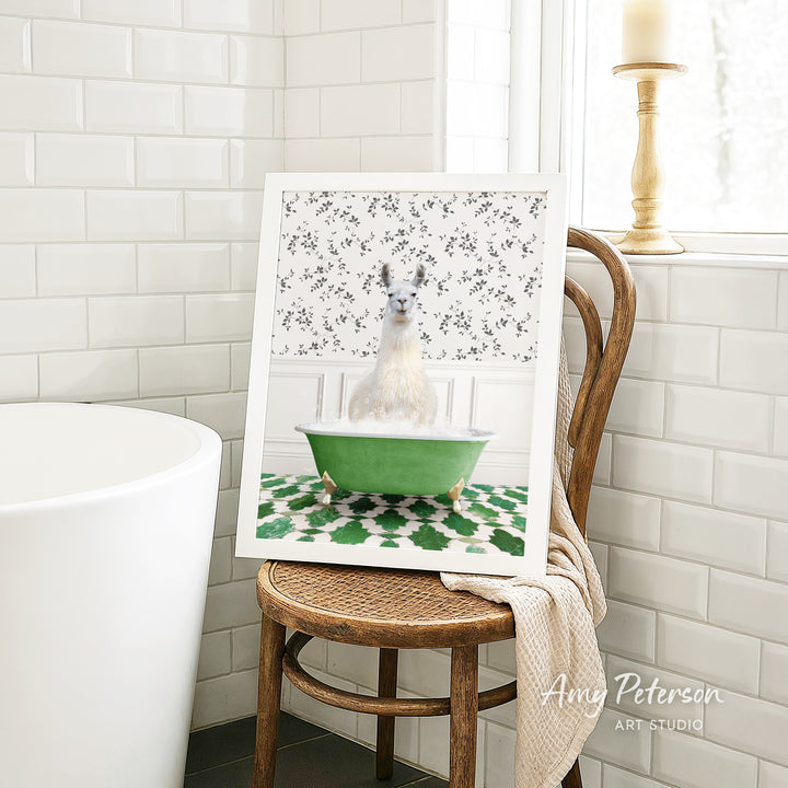 A framed image of a white llama sitting in a green bathtub, surrounded by a patterned design, is displayed on a wooden chair in a bathroom setting.