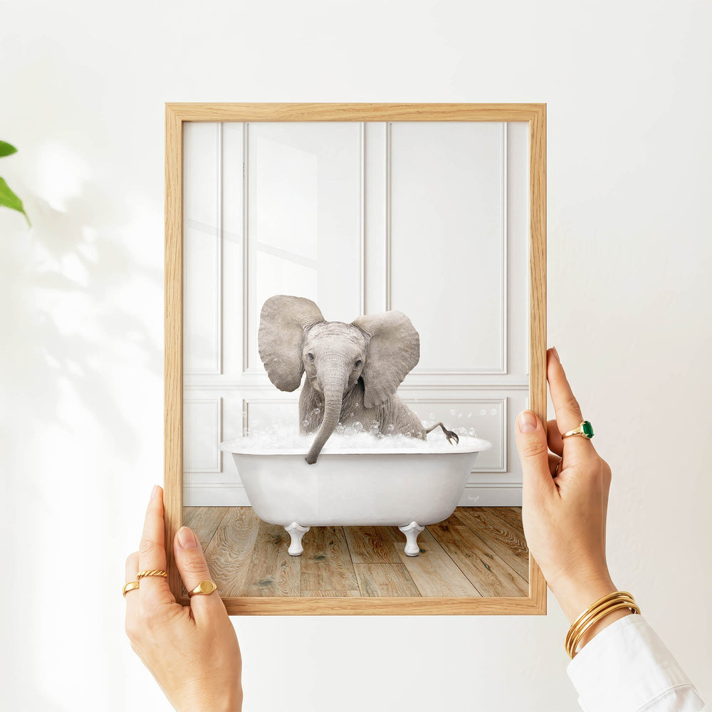 A person's hands are holding a picture frame with a photograph of a baby elephant sitting in a bathtub.