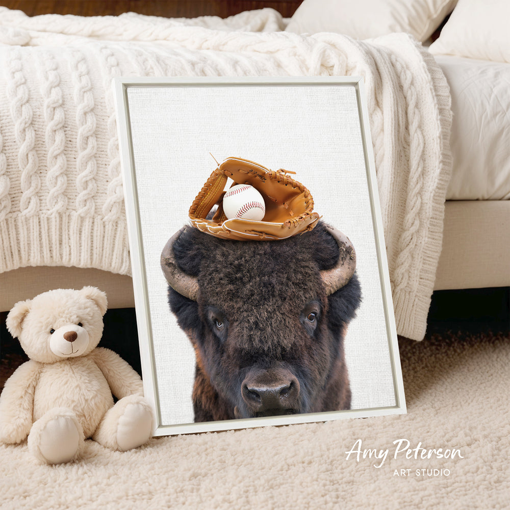 A framed print of a buffalo's head with a baseball glove on top, placed on a bed next to a stuffed teddy bear.
