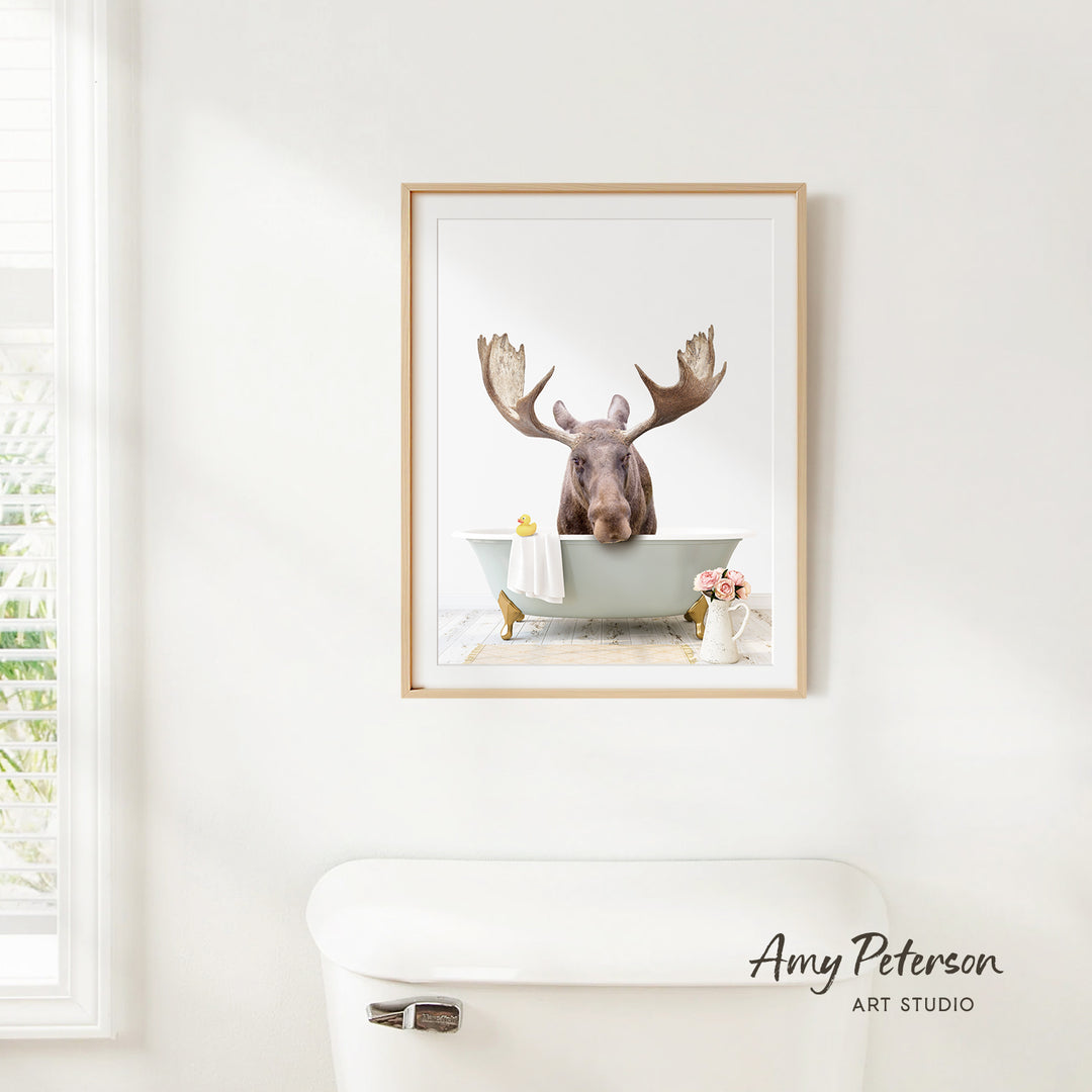 A framed image of a moose sitting in a bathtub, with a vase of flowers and a white toilet in the foreground.