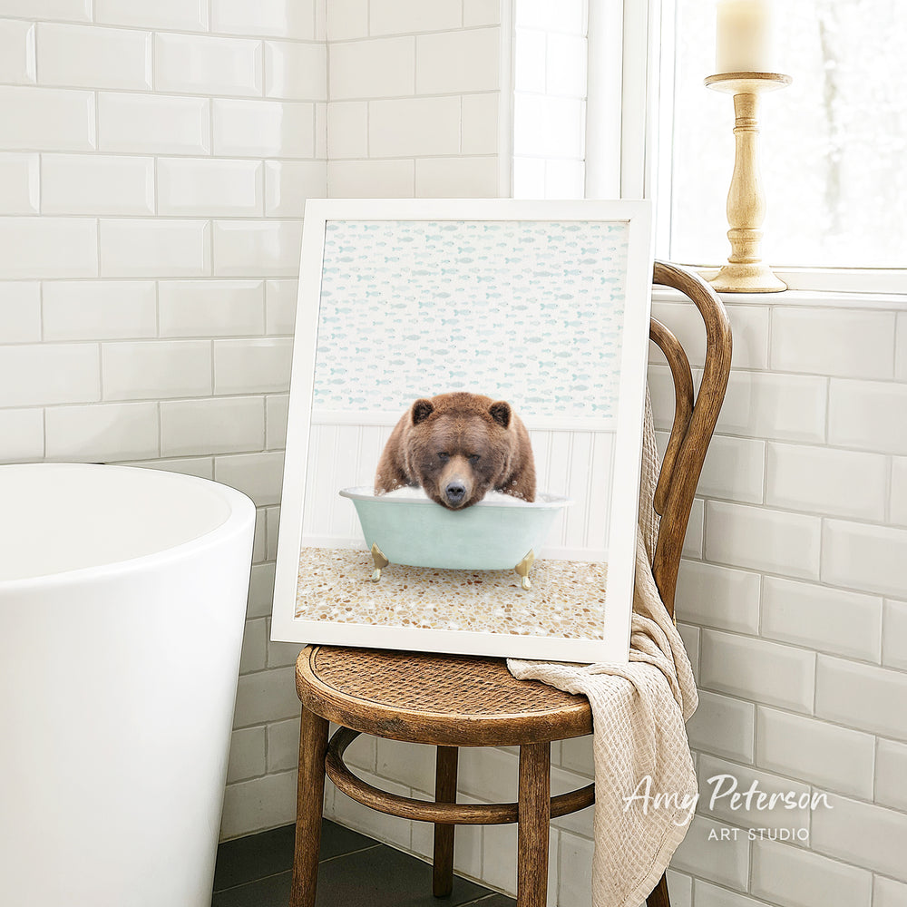 A framed image of a brown bear sitting in a bathtub, with a towel draped over the back of a wooden chair in a bathroom setting.