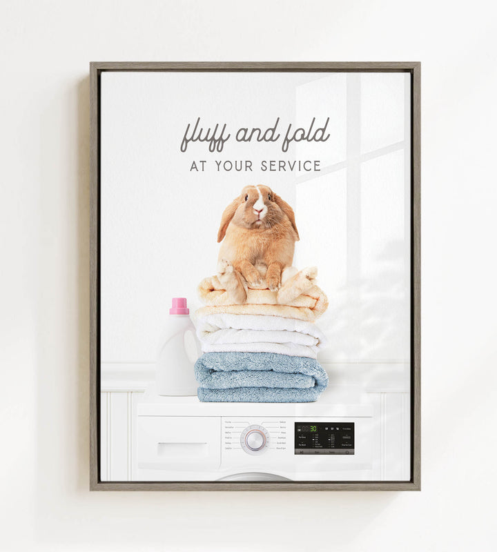 A framed image of a rabbit sitting on top of a stack of folded towels, with the text "fluff and fold at your service" above the rabbit.