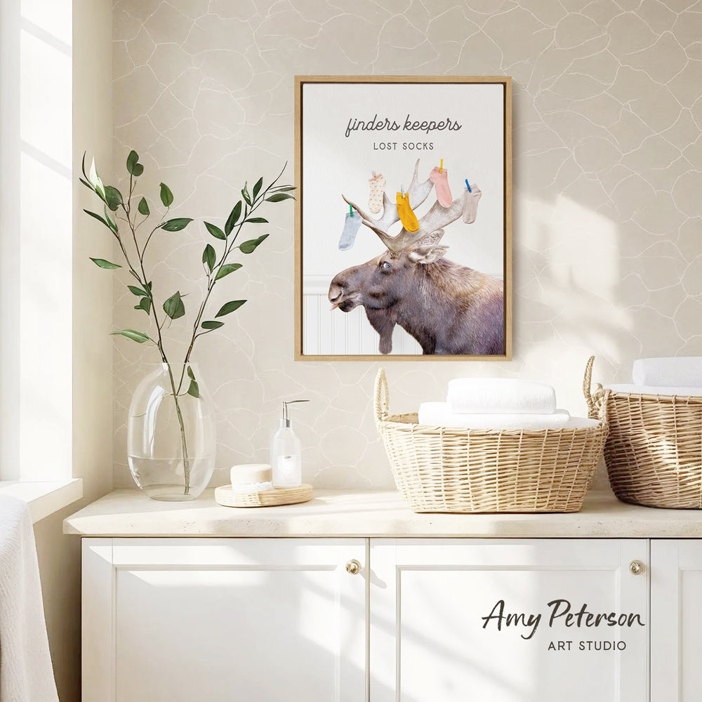 a framed poster of a moose with a sign that says "finder's keepers" above it, placed on a wall above a white cabinet. The cabinet has two woven baskets on top of it, and there is a vase with a plant on the left side of the cabinet.