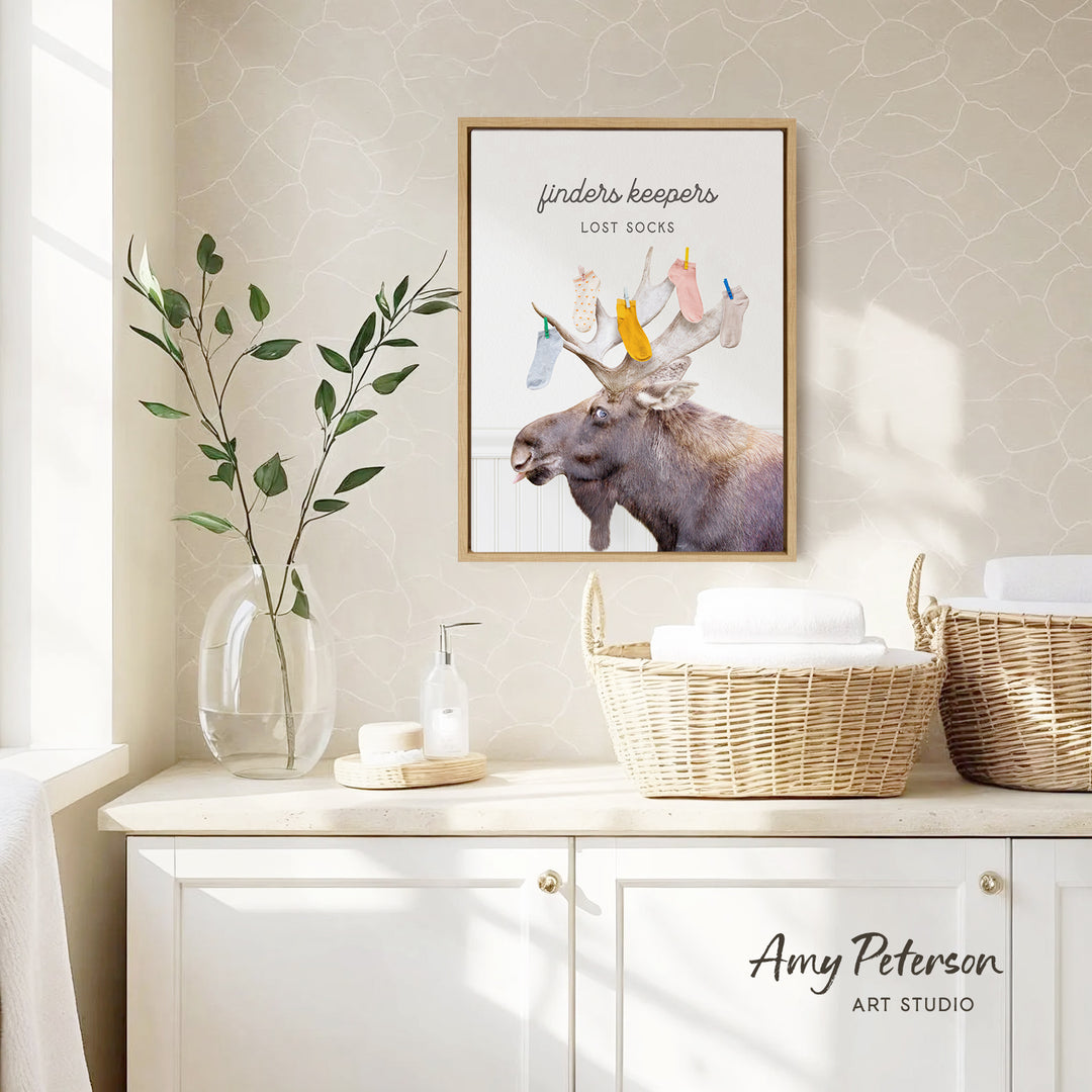 a framed poster of a moose with a sign that says "finder's keepers" above it, placed on a wall above a white cabinet. The cabinet has two woven baskets on top of it, and there is a vase with a plant on the left side of the cabinet.