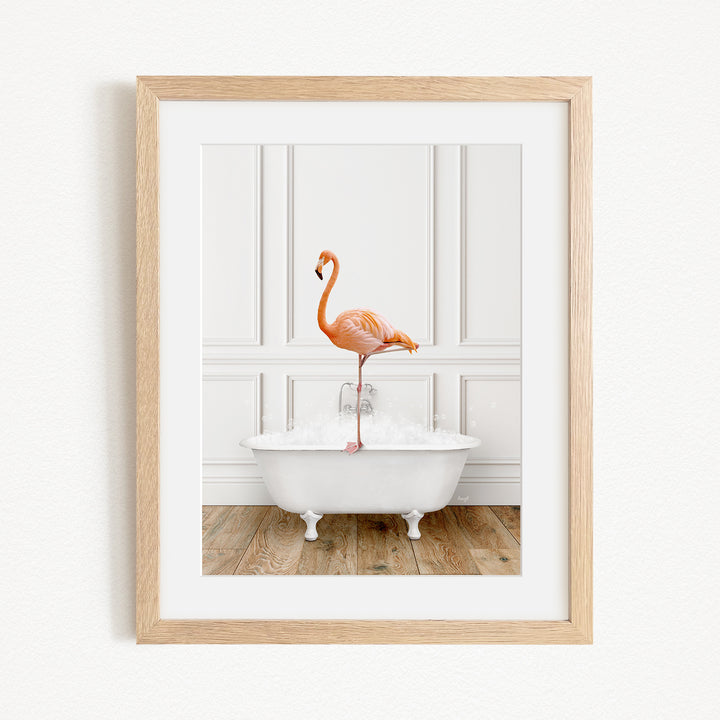 A framed image of a pink flamingo standing on the edge of a white clawfoot bathtub, with a wooden floor and white walls in the background.