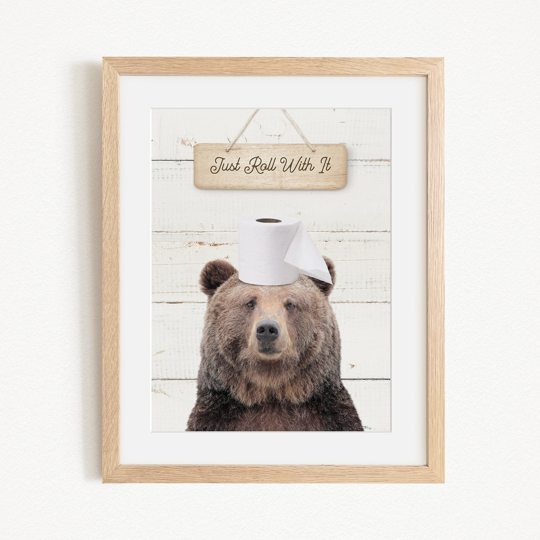 A framed image of a brown bear wearing a toilet paper hat, with a wooden sign above it that says "Just Roll With It".