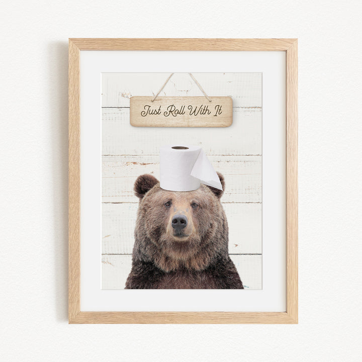 A framed image of a brown bear wearing a toilet paper hat, with a wooden sign above it that says "Just Roll With It".