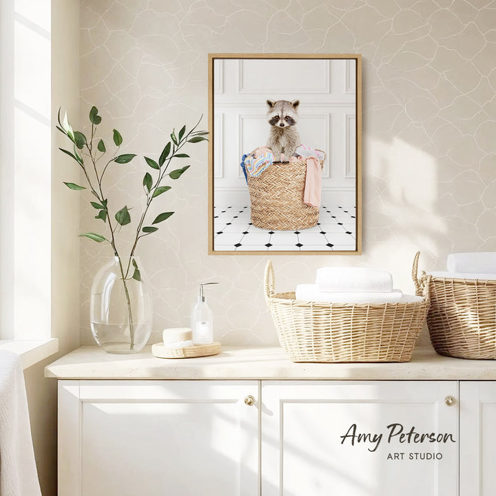 A framed photograph of a raccoon sitting in a woven basket is displayed on a wall in a bathroom setting, with a vase of leaves and a bottle of soap nearby.