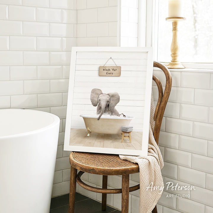 A framed image of an elephant sitting in a bathtub, with a sign above it that says "Hush Your Ear". The image is displayed on a wooden chair in a bathroom setting.