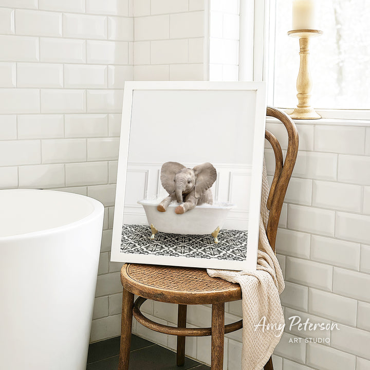 A framed image of a baby elephant sitting in a bathtub, with a towel draped over the back of a wooden chair in a bathroom setting.