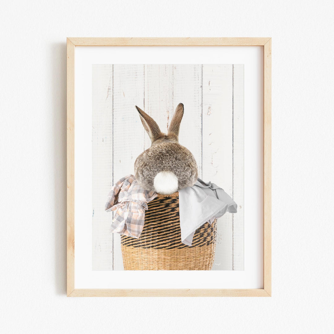 A gray rabbit sits in a wicker basket filled with clothes, with its back turned to the viewer.
