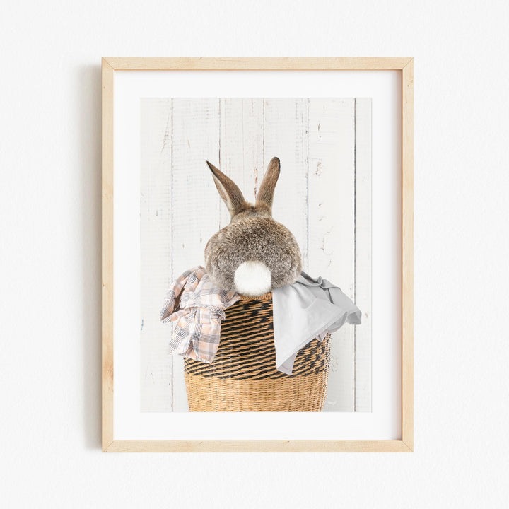 A gray rabbit sits in a wicker basket filled with clothes, with its back turned to the viewer.