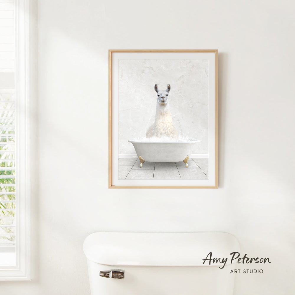 A framed image of a white llama sitting in a white bathtub is hanging on a white wall above a white toilet.