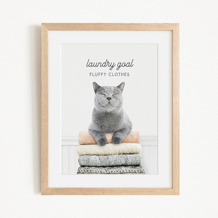 A framed image of a gray cat sitting on a stack of fluffy clothes, with the text "laundry goal" and "fluffy clothes" written above it.
