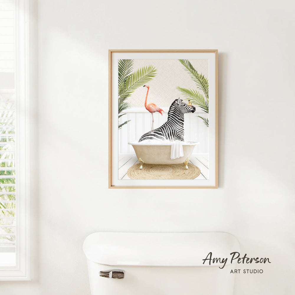A framed print of a zebra sitting in a bathtub, surrounded by a pink flamingo and palm leaves, hangs on a white wall.