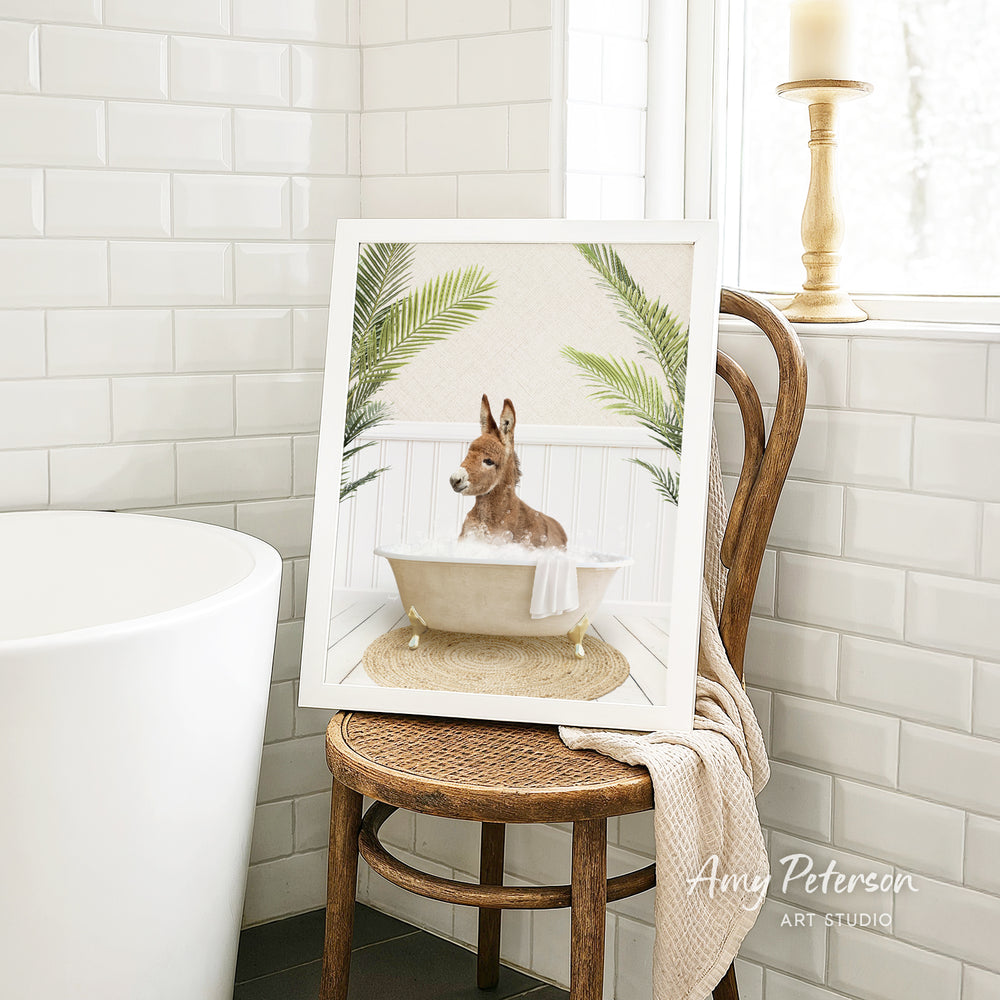A framed image of a dog sitting in a bathtub, surrounded by palm leaves, is displayed on a wooden chair in a bathroom setting.