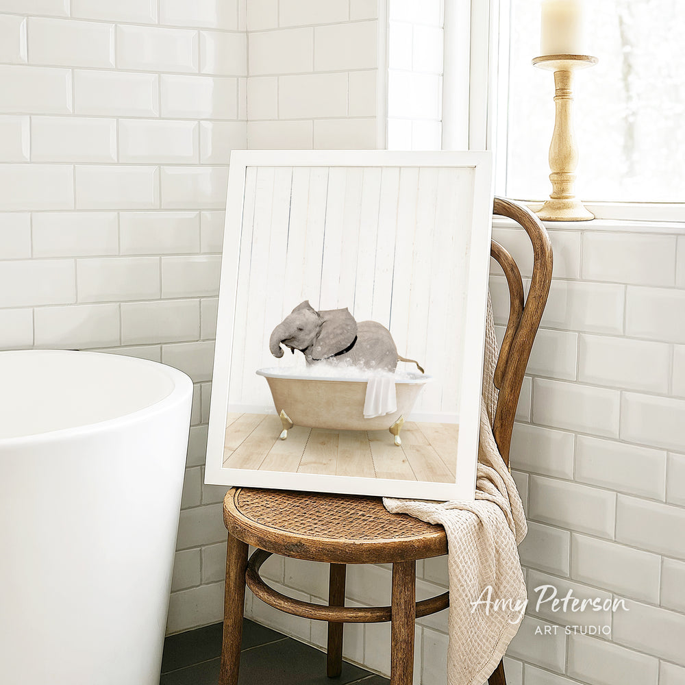 A framed image of a baby elephant sitting in a bathtub, surrounded by white tiles, is displayed on a wooden chair in a bathroom setting.