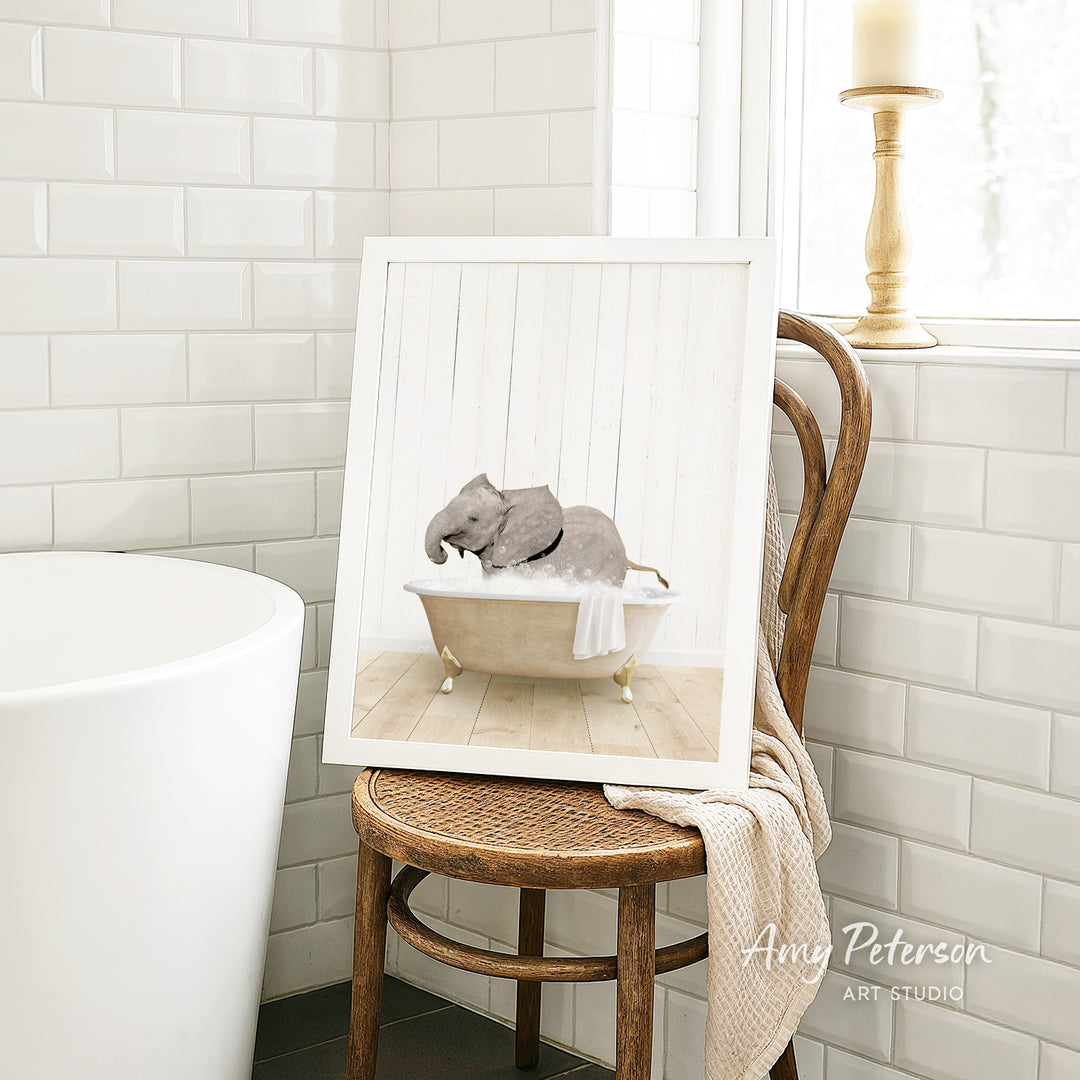 A framed image of a baby elephant sitting in a bathtub, surrounded by white tiles, is displayed on a wooden chair in a bathroom setting.