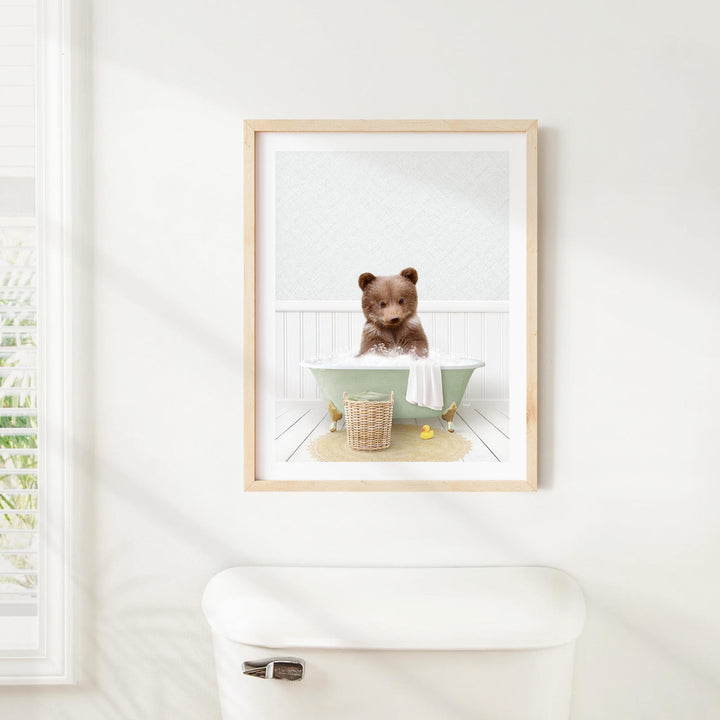 A framed image of a brown bear sitting in a bathtub, surrounded by a basket of bath toys and a towel, is displayed on a white wall above a white toilet.