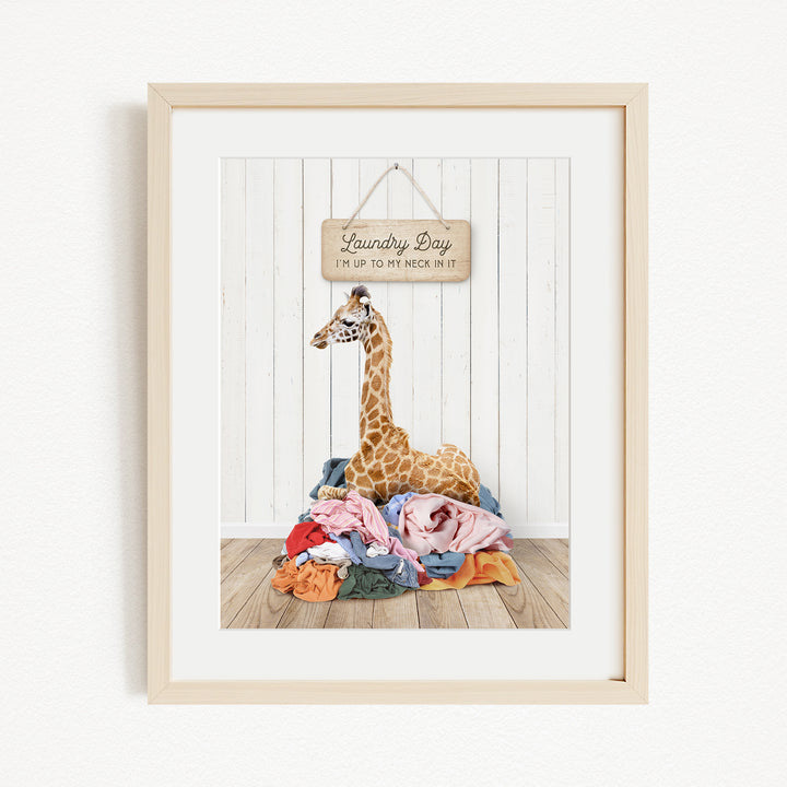 A giraffe is sitting on a pile of clothes in front of a wooden sign that says "Laundry Day".