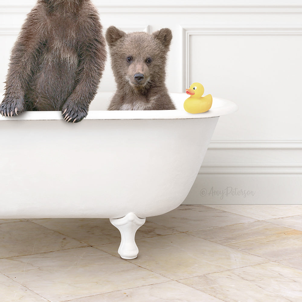 A baby bear and a rubber duck are sitting in a white bathtub, with the bear looking at the camera.