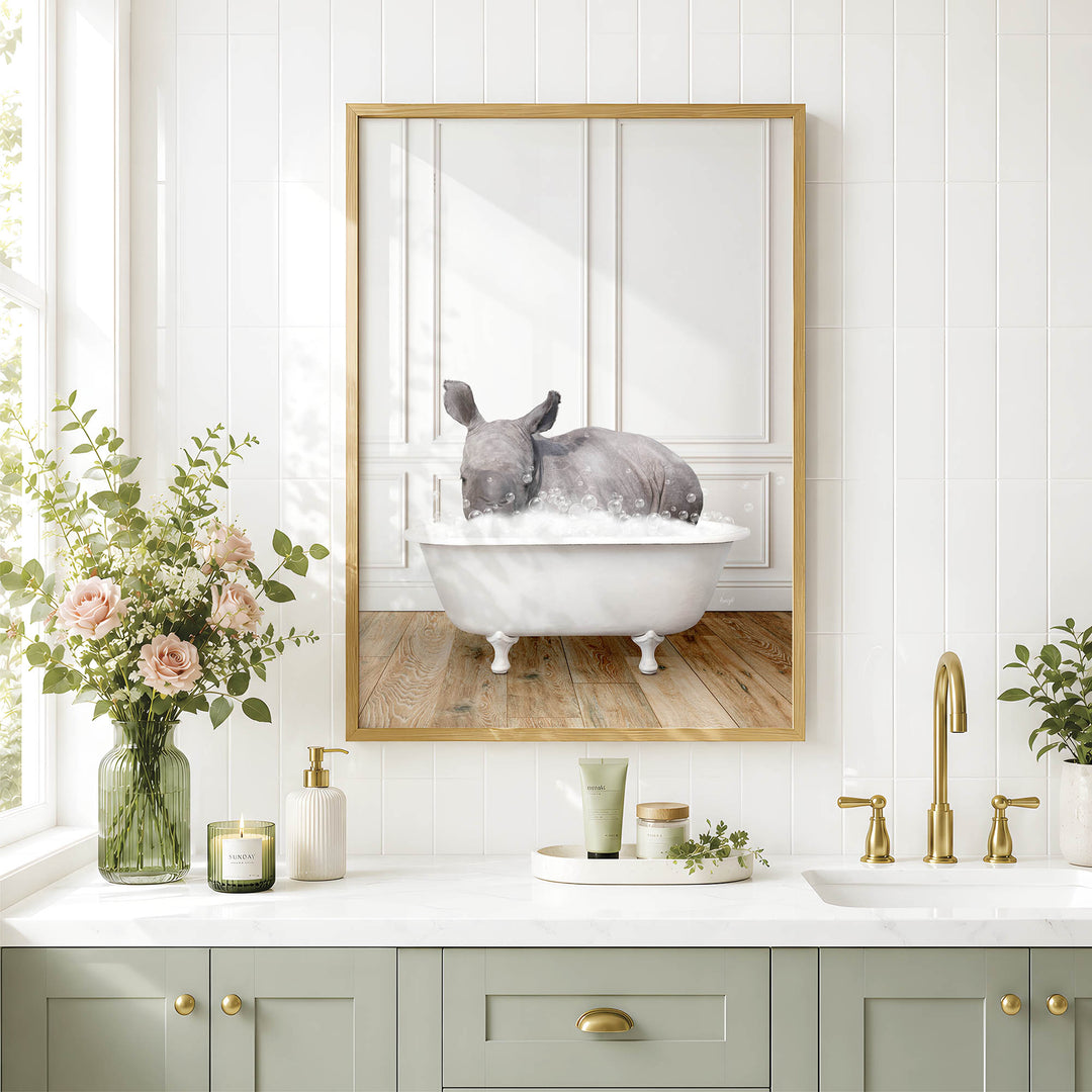 A framed image of a rhinoceros sitting in a bathtub, surrounded by a vase of flowers, candles, and a green plant on a white countertop in a bathroom setting.