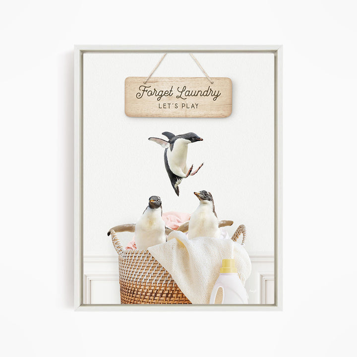 A framed image depicting three penguins in a basket filled with laundry, with a sign above that says "Forget Laundry, Let's Play".