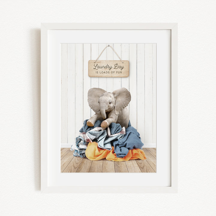 A framed image of an elephant sitting on a pile of laundry, with a sign above that says "Laundry Day is loads of fun".