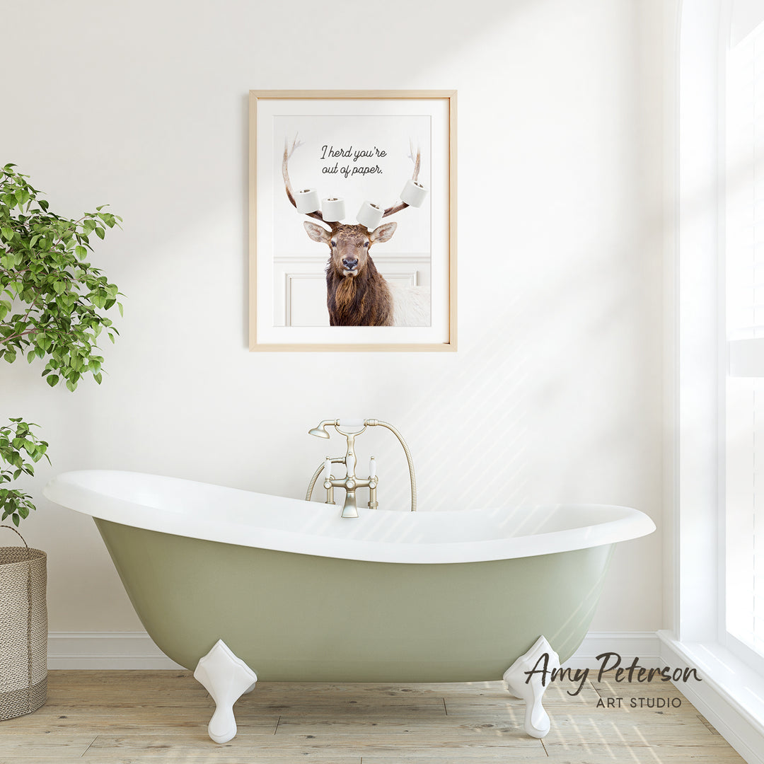 A framed image of a deer with antlers is displayed on the wall above a green clawfoot bathtub in a bright, white bathroom.