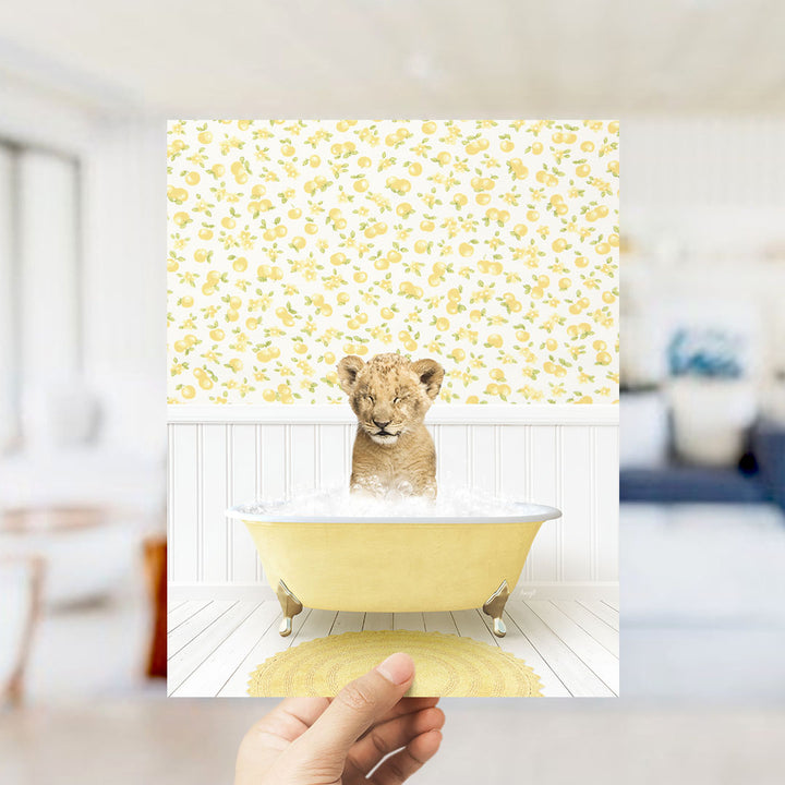 A hand holds up a framed image depicting a young lion cub sitting in a yellow bathtub filled with water, surrounded by a yellow and white floral pattern.