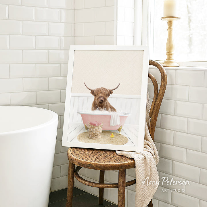 A framed image of a bulldog sitting in a pink bathtub, with a towel and a basket of toys nearby, is displayed on a wooden chair in a bathroom setting.