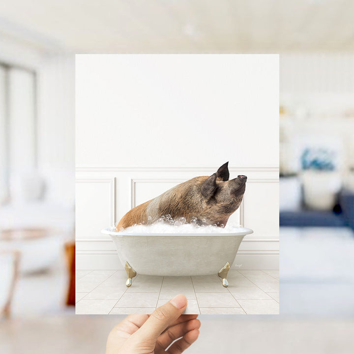 A hand holds up a framed image of a pig sitting in a bathtub filled with bubbles.