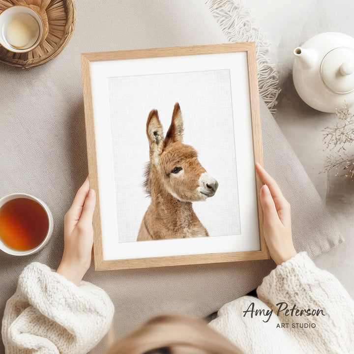 A person is holding a framed picture of a donkey with a teacup and saucer nearby, and a teapot on a table.